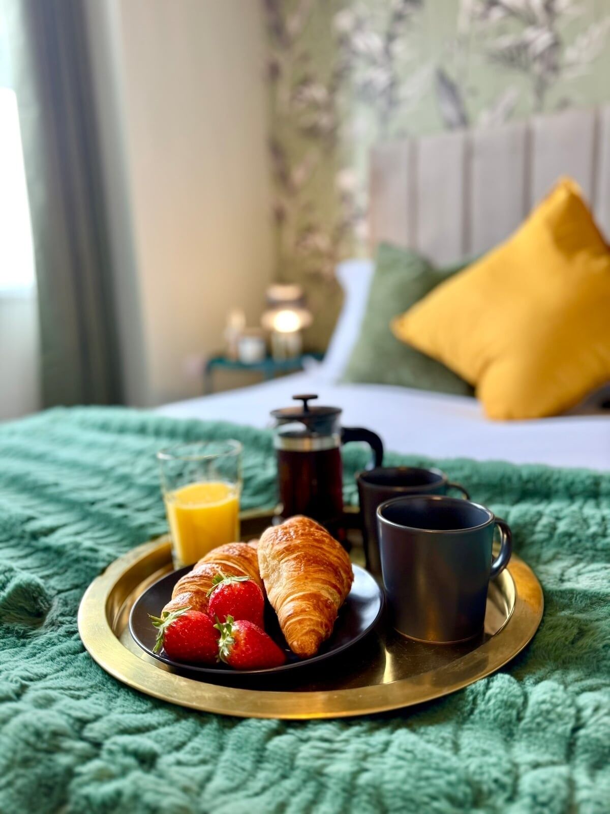 Een ontbijfwagen met croissants, aardbeien en een glas sinaasappelsap op een bed met een groene deken en een geel kussen.