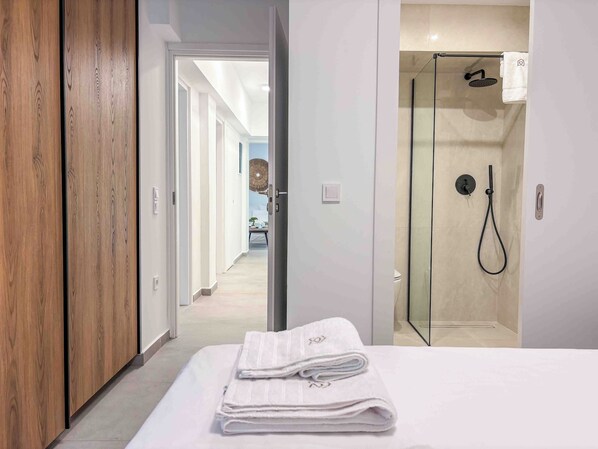 A hotel room with a wooden wardrobe, a bed with neatly folded towels, and a glass-enclosed shower area. - Elegant Flat in the Center of Glyfada (Glifada)