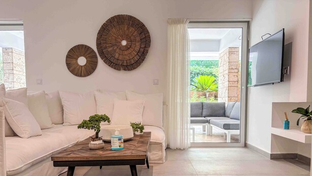 A modern living room with a white sofa, a coffee table, a TV mounted on the wall, and a view of the outdoors through a glass door. - Elegant Flat in the Center of Glyfada (Glifada)