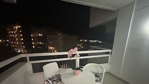 Un balcon avec une table blanche et deux chaises, un vase rose contenant une fleur et une balustrade.