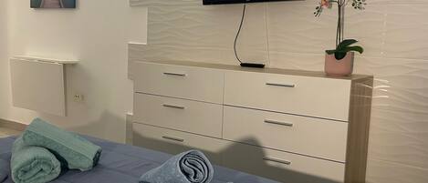 A bedroom with a bed, a TV mounted on the wall, a dresser, a painting, and a potted plant.