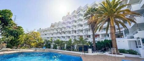 A swimming pool with blue tiles, surrounded by palm trees and a white building with balconies.