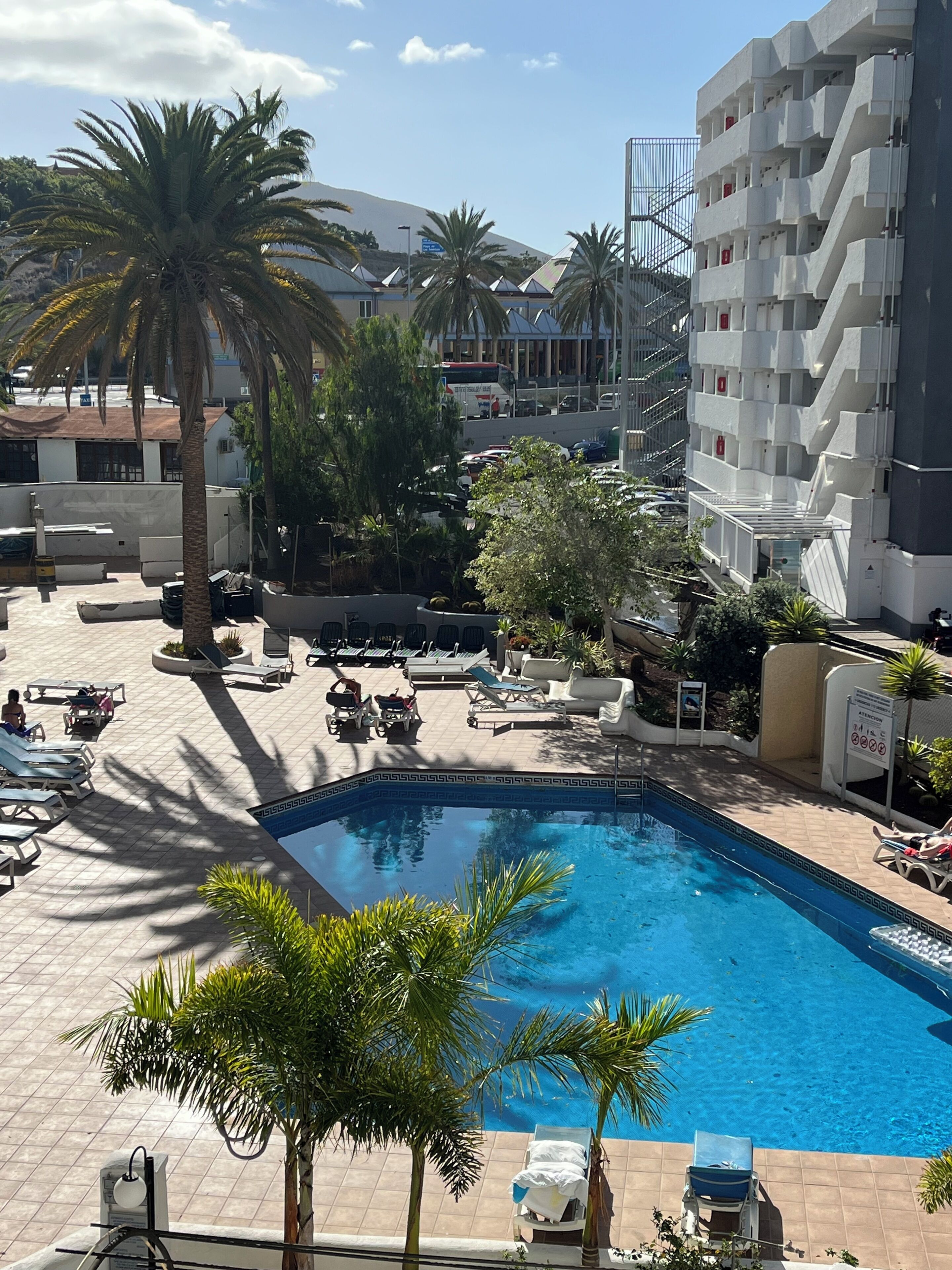 A swimming pool area with lounge chairs and palm trees, surrounded by buildings.