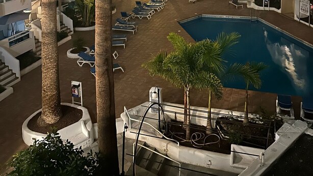 Un espace piscine aménagé avec des chaises longues, des palmiers et une piscine entourée de bâtiments.