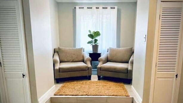 Un petit espace salon fermé, comprenant deux canapés et un tapis, une plante en pot et une fenêtre avec des stores.