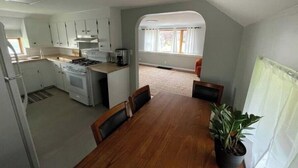 A compact kitchen with white cabinets, a stove, and a dining area with a wooden table and chairs. - Cozy Cuddler's Cottage (West Falls)
