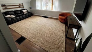 A compact living space with a black leather sofa, a beige rug, a wooden coffee table, a small orange armchair, and a flat-screen TV mounted on the wall. - Cozy Cuddler's Cottage (West Falls)