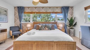 A spacious bedroom with a large bed, a ceiling fan, and a view of palm trees through the windows.