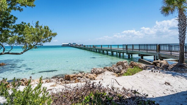 A beach with clear turquoise water, a wooden pier extending into the sea, and a sandy shore with sparse vegetation.