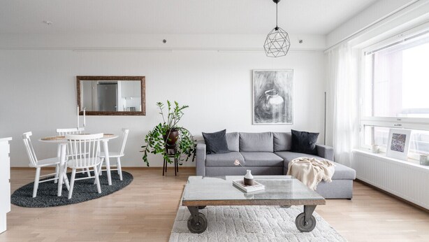 A modern living room with a grey sofa, a round dining table with chairs, a coffee table, a potted plant, and a large window with a view.