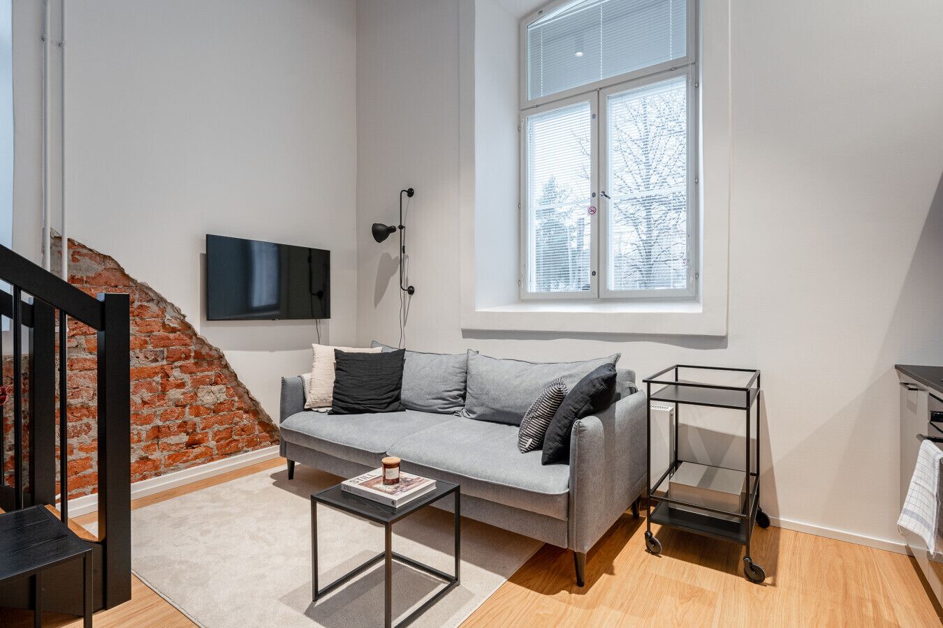 A modern living room with a grey sofa, a flat-screen TV mounted on the wall, a window with blinds, a small table, and a black metal cart.