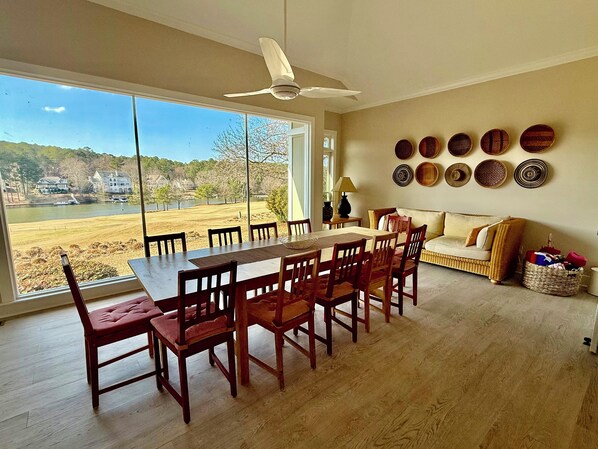 A dining area with a large table and chairs, a sofa, and a view of a lake and houses.