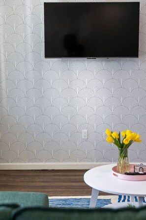 Une pièce avec un papier peint à motifs, un téléviseur fixé au mur, une petite table ronde ornée d’un vase de fleurs jaunes et un canapé vert.