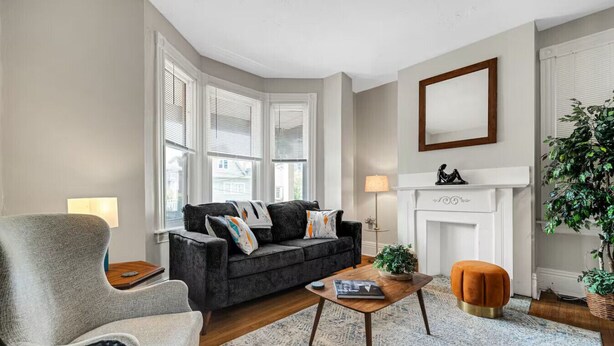 A living room with a dark grey sofa, a light grey armchair, a wooden coffee table, and a small orange ottoman. There is a fireplace with a white mantel and a mirror above it. - 5BDRM GEM with Arcade - Mt Wash (Pittsburgh)