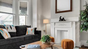 A modern living room with a dark grey sofa, a wooden coffee table, and an orange ottoman. There is a fireplace with a decorative mantel and a mirror above it. - 5BDRM GEM with Arcade - Mt Wash (Pittsburgh)