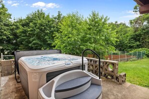 A hot tub with a built-in seat, surrounded by a concrete deck, with a wooden fence and lush greenery in the background.