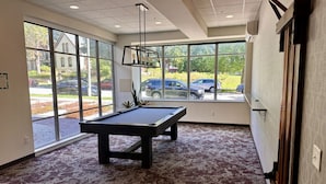 Mesa de billar en una habitación con ventanas amplias, piso alfombrado y vista al exterior con coches estacionados y vegetación.