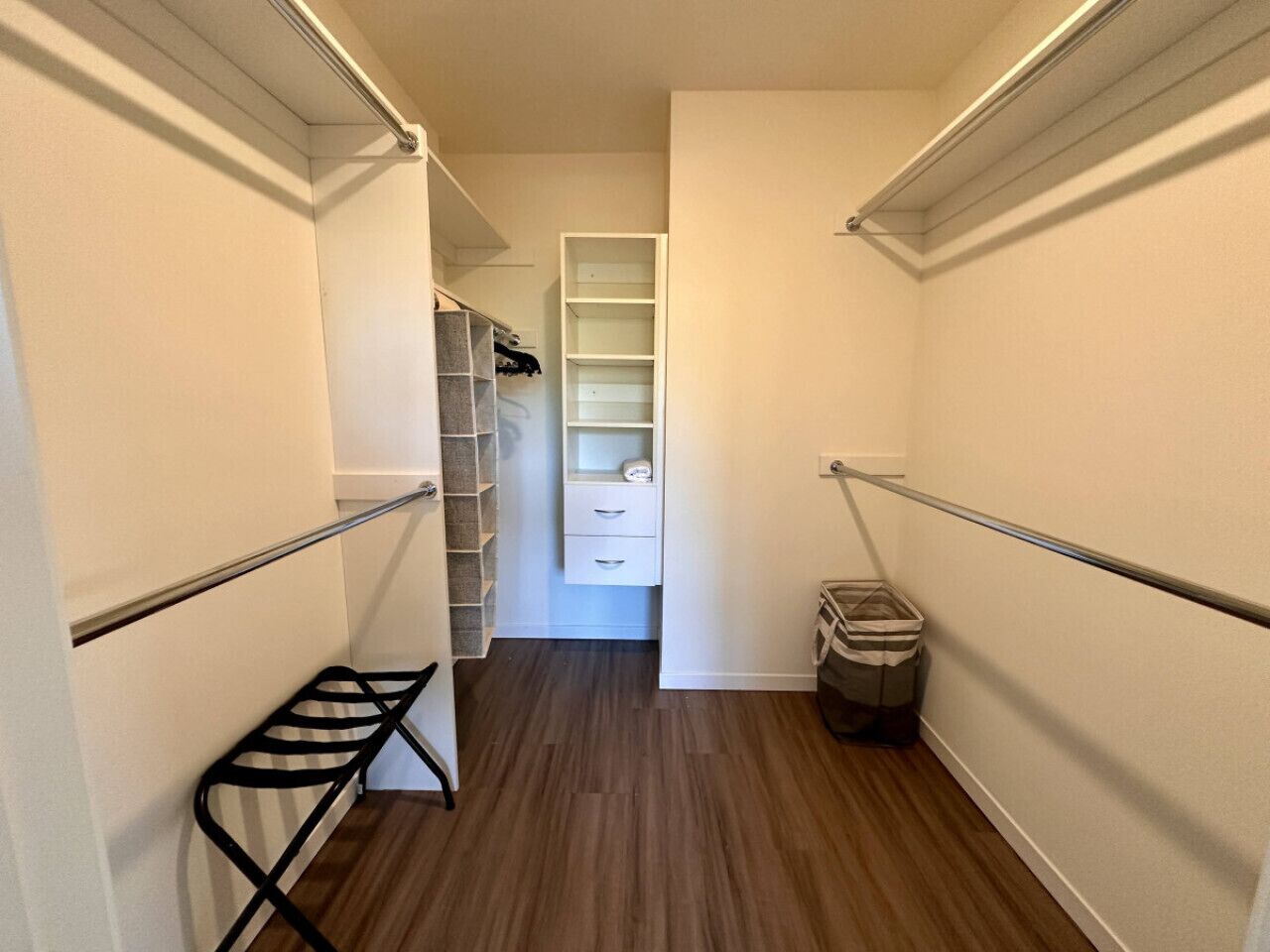 Un pasillo estrecho con un tendedero de metal, un bote de basura y un mueble blanco con cajones.