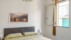 Et soveværelse med en seng, et natbord, en lampe, et maleri af en solnedgang og et vindue med gardiner.