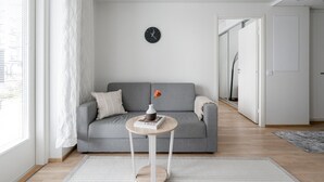 Ein modernes Wohnzimmer mit einem grauen Sofa, einem kleinen runden Tisch mit Büchern, einer Wanduhr und Blick auf eine Stadtlandschaft durch das Fenster.