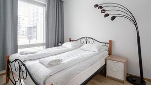 Ein ordentlich bezogenes Bett mit weißen Leinen, ein Nachttisch mit Schublade, eine Stehlampe und ein Fenster mit Vorhängen.