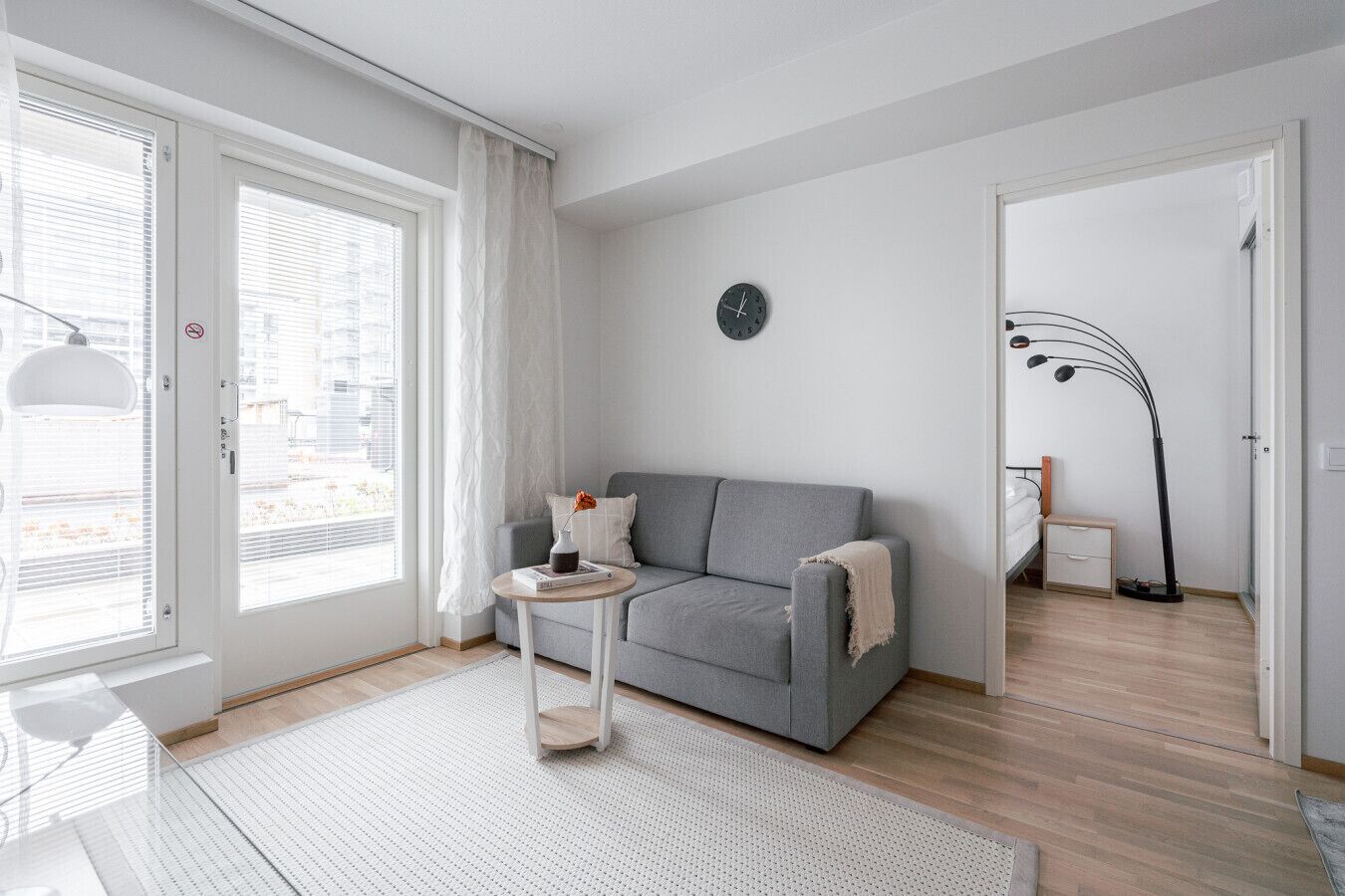Een moderne woonkamer met een grijze bank, een klein rond tafeltje, een vloerlamp en uitzicht door een raam met jaloezieën.