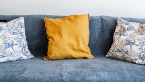 Een blauwe bank met een mosterdkleurig kussen en twee gestreepte kussens, een luchtverfrisser aan de muur en vier geweven wanddecoraties.