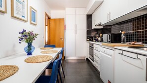 Een praktische keuken met witte kastjes, een zwart betegelde spatwand en een eethoek met een witte tafel en blauwe stoelen.