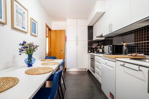 Una cucina compatta con mobili bianchi, uno schienale nero in piastrelle e un'area pranzo con tavolo bianco e sedie blu.