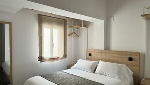 Una cama bien hecha con cabecera de madera, un perchero y una ventana con cortinas de gasa.