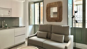 Una cocina moderna con armarios blancos, horno empotrado y pileta. Hay un sofá color beige con cojines y un alfombra redonda en el piso. Un espejo grande cuelga de la pared, y se ve una lámpara de pie.