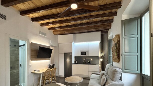 Un dormitorio moderno con una cama grande, un pequeño comedor, una kitchenette y una sala de estar con sofá y televisor.