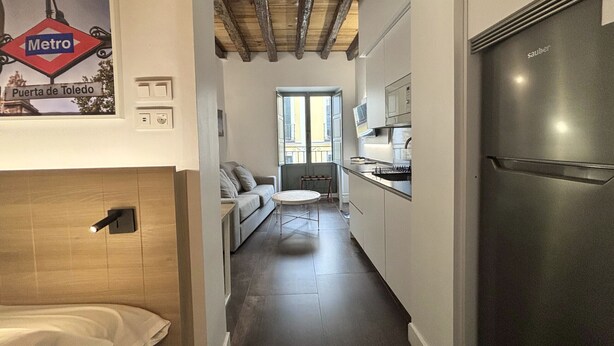 Un pasillo estrecho que da a una cocina con un refrigerador moderno de acero inoxidable, un sofá y un ventanal con vistas al exterior.