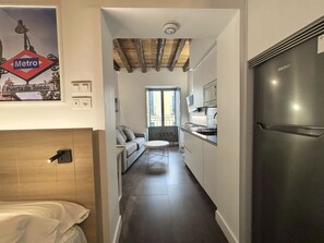 A narrow hallway leading to a kitchen with a modern stainless steel refrigerator, a sofa, and a window with a view of the outside.