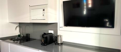A modern kitchen with white cabinets, a built-in microwave, a coffee machine, and a large flat-screen TV mounted on the wall.