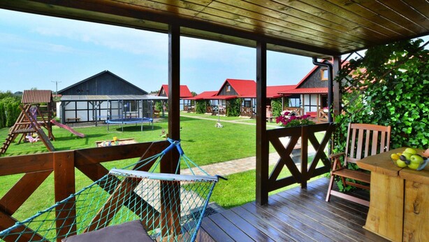 A wooden deck with a hammock, a wooden table with fruit, and a view of a green lawn and buildings in the background. - Cottage in Bobolin Near Baltic Beach (Darlowo)
