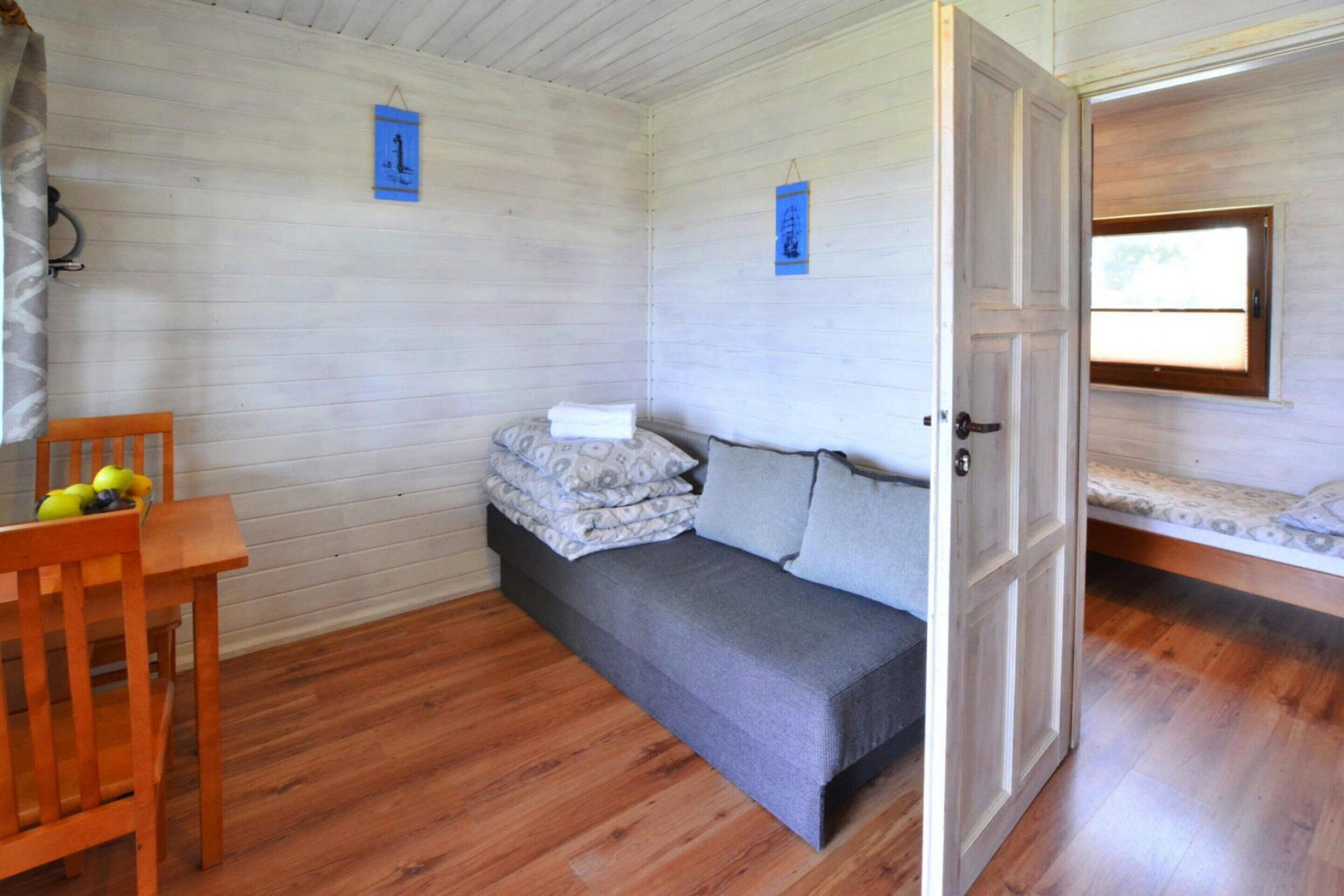 A room with a wooden floor, a bed with a grey mattress and pillows, a wooden chair with yellow objects, and a door leading to another room.