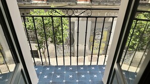 A balcony with a blue tiled floor featuring white stars, a metal railing, and a view of the street with closed shops and greenery.