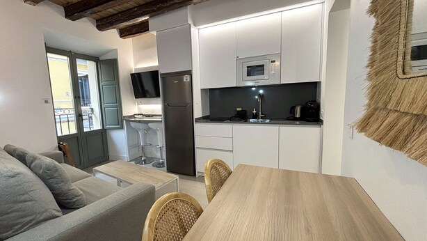 Una cocina compacta con armarios blancos, un refrigerador de acero inoxidable, un microondas y un lavamanos. Hay una mesa de comedor de madera con sillas y un sofá gris en la zona de estar contigua.