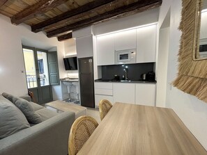 Eine kompakte Küche mit weißen Schränken, einem Edelstahlkühlschrank, einer Mikrowelle und einem Spülbecken. Im angrenzenden Wohnbereich befindet sich ein Holzessttisch mit Stühlen und ein graues Sofa.