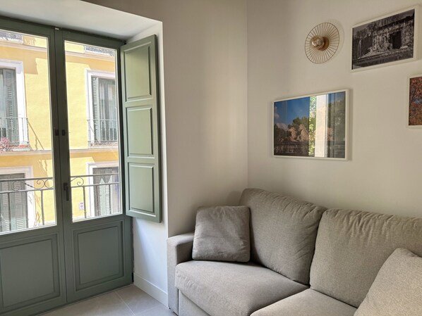 Una habitación con un sofá, una ventana con contraventanas abiertas y dos cuadros en la pared.