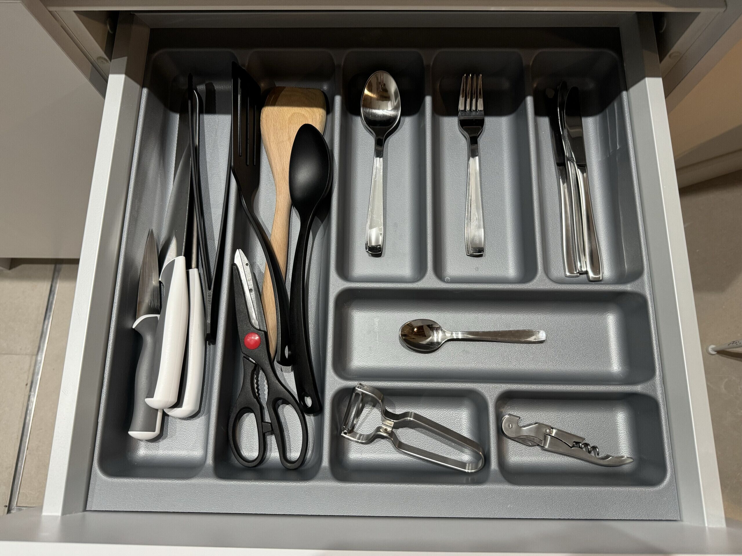 A drawer with compartments containing various kitchen utensils such as knives, spoons, forks, and scissors.