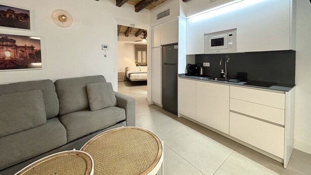 A modern kitchen with white cabinets, a microwave, and a sink. There is a gray sofa and a wicker chair in the living area.