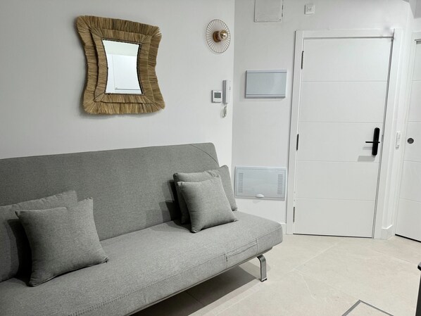 A modern living room with a grey sofa, two cushions, a decorative mirror, a wall-mounted thermostat, and a white door.