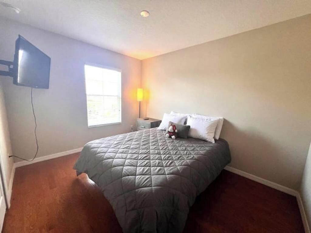 A bedroom with a bed, a flat-screen TV mounted on the wall, a window with a view, and a nightstand with a lamp.