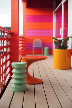 A wooden deck with a red and orange striped wall, a small orange table, a stack of green stools, and a potted plant. - Hotel Hilo (Louisville)