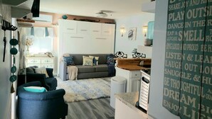 Una sala de estar con un sofá gris, un sillón azul y una mesa de centro. Hay una zona de cocina con un lavamanos y una encimera.