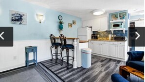 Cocina compacta con armarios blancos, microondas y tostadora. Hay dos taburetes de bar y una mesita pequeña. Las paredes están pintadas de azul claro y hay un ventilador de techo.