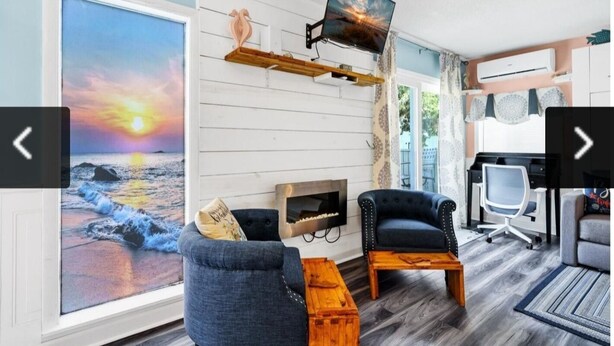 Un salón con un ventanal grande, una chimenea, dos sillones, una mesa de centro de madera, un televisor montado en un estante de madera y un escritorio con una silla.