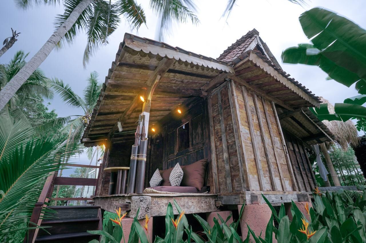 A rustic wooden structure with a thatched roof, a porch with cushions, and a staircase leading up.